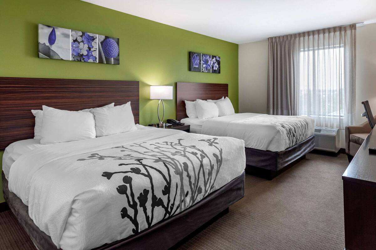 Quarto de hotel com duas camas, uma escrivaninha e uma cadeira. Há quadros nas paredes acima das camas e uma janela grande com cortinas.