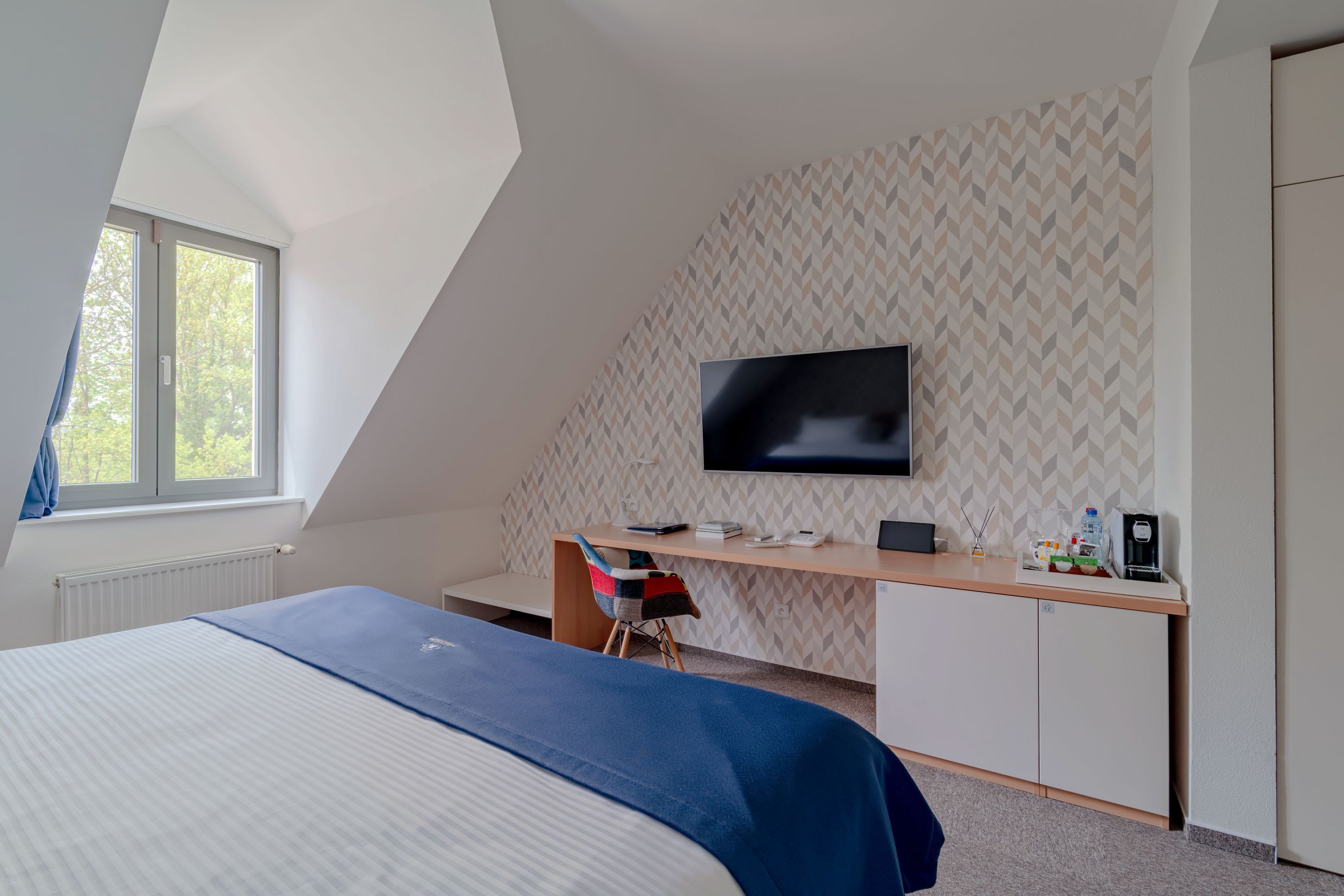 A bedroom with a bed, a desk, a chair, a television, and a window.
