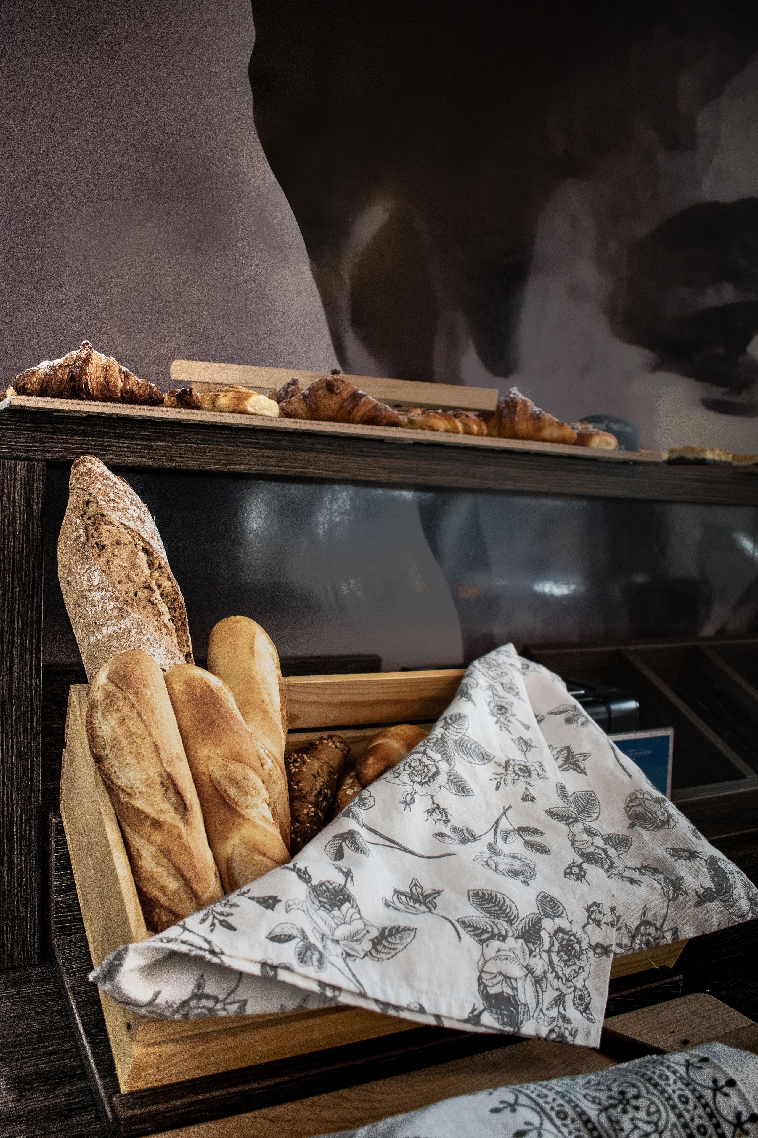 Ein Holzkörbchen mit verschiedenen Brotsorten und einer Blumen-Serviette auf einem Tisch.