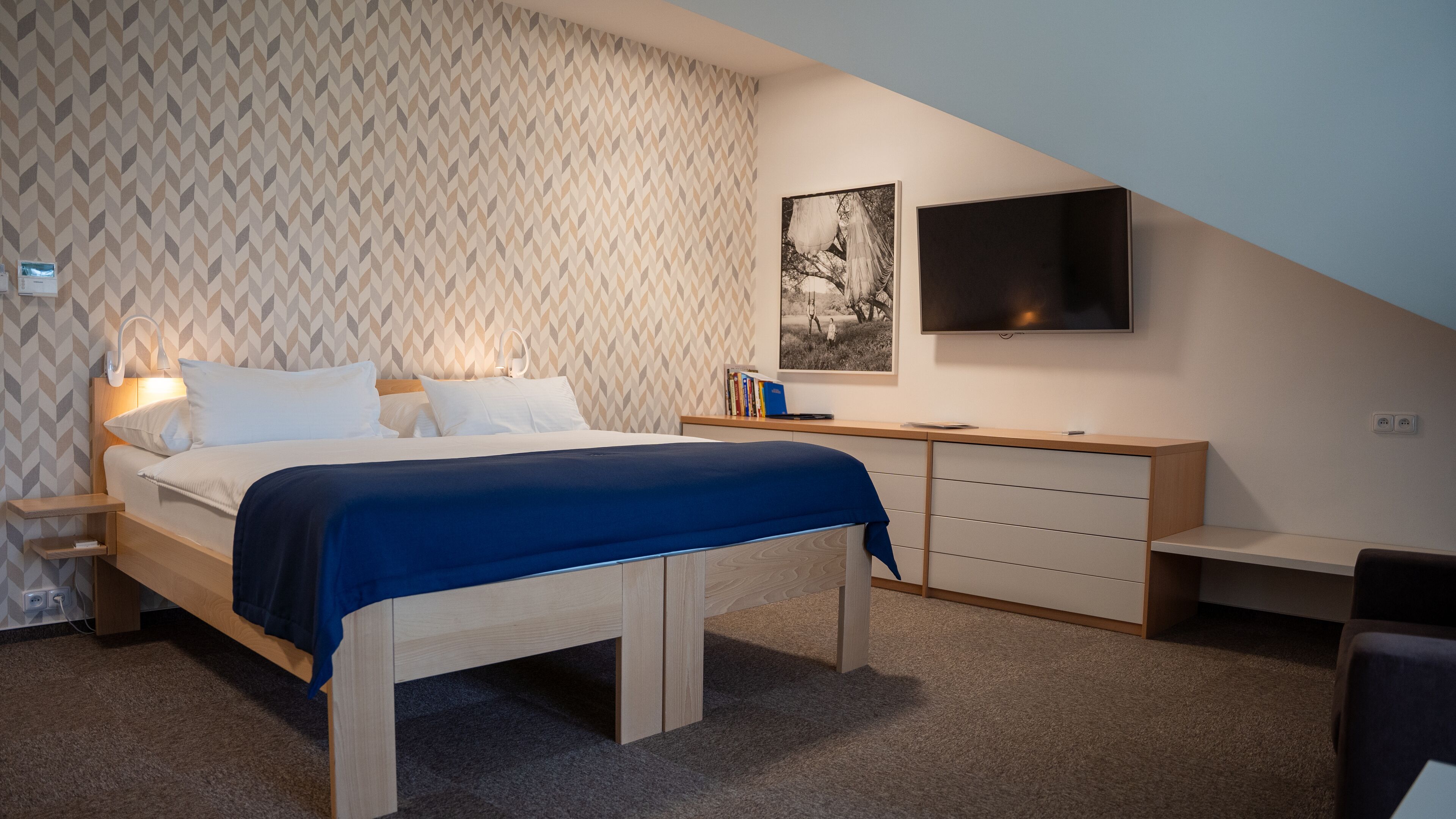 A bedroom with a bed, bedside tables, a desk, a chair, a TV, and a patterned wall.