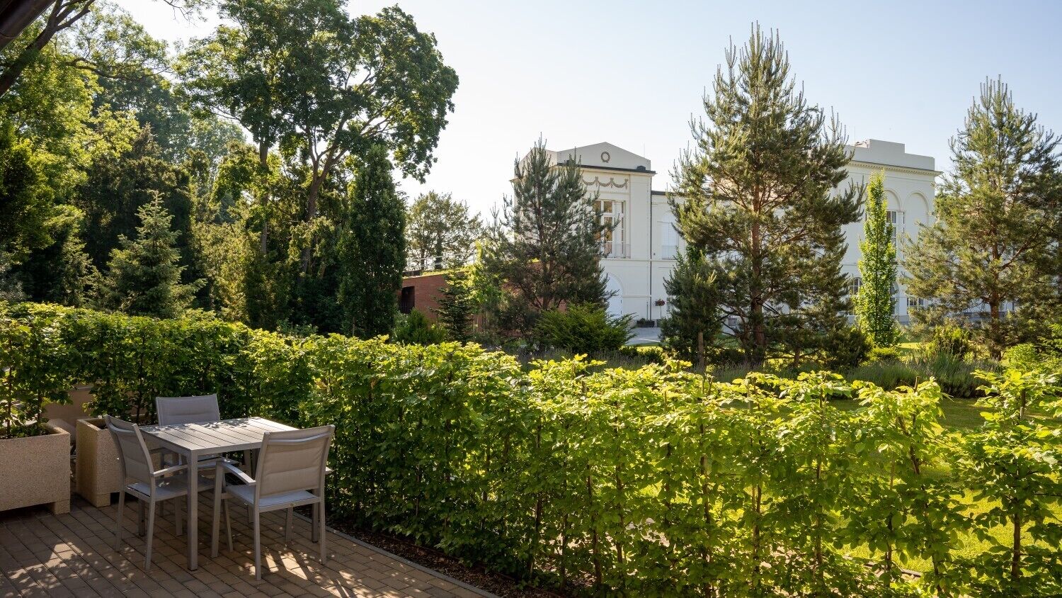 A patio with a table and chairs, surrounded by lush greenery and a white building in the background.