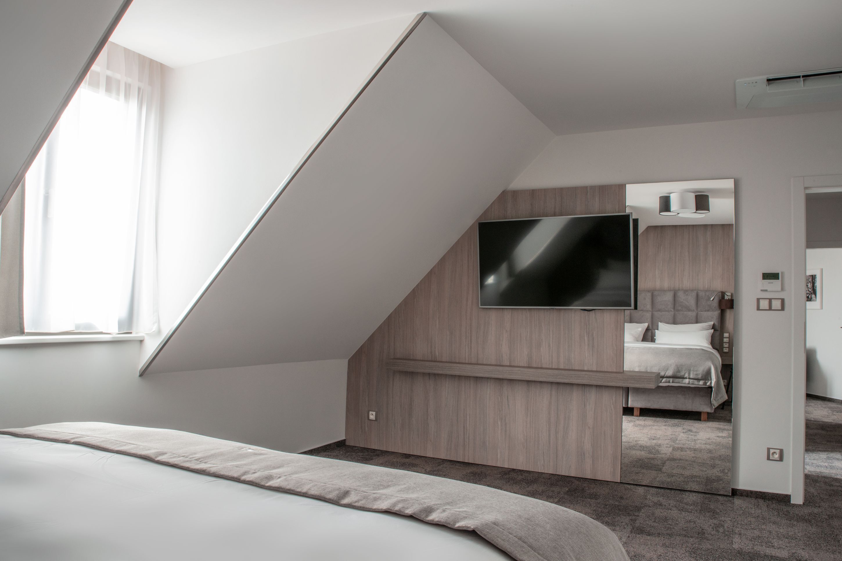 A modern bedroom with a sloped ceiling, a large bed, a TV mounted on the wall, and a visible door leading to another room.