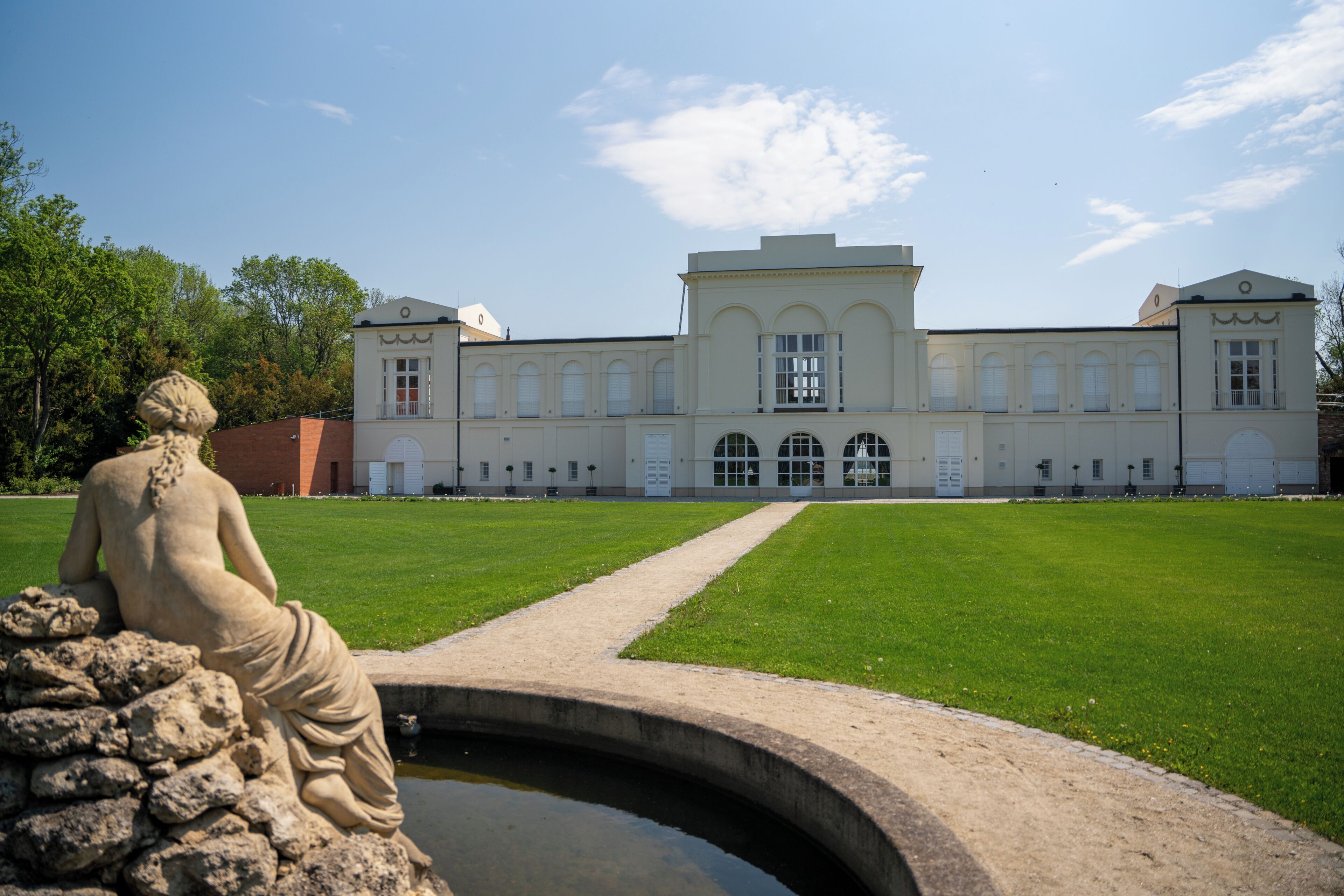 Un bâtiment de style classique, doté d’une fenêtre en arc central, d’une statue et d’un puits en premier plan.