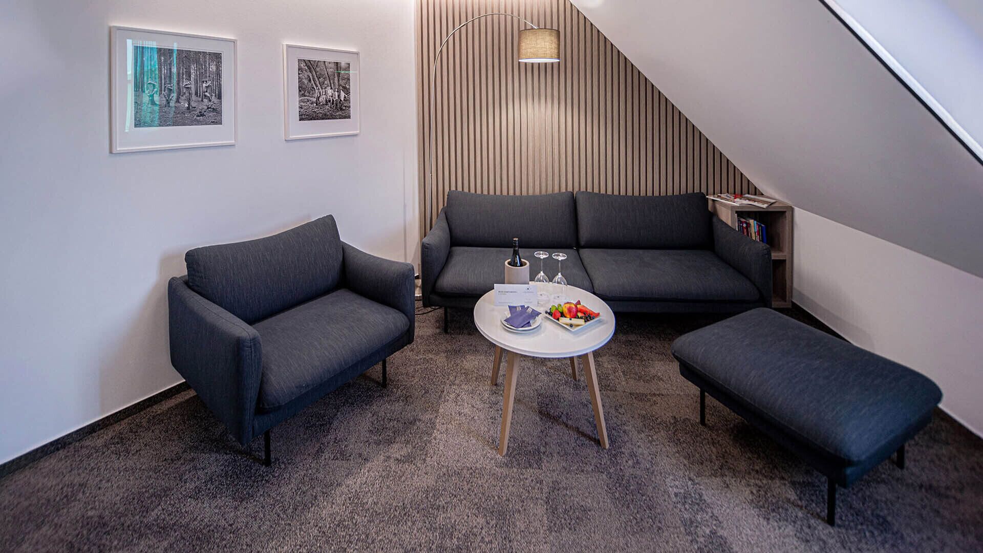 Una sala de estar moderna con un sofá gris, dos sillones, una mesa de centro con fruta y copas de vino, y una escalera que conduce a otro nivel.
