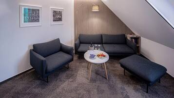 Una sala de estar moderna con un sofá gris, dos sillones, una mesa de centro con fruta y copas de vino, y una escalera que conduce a otro nivel.