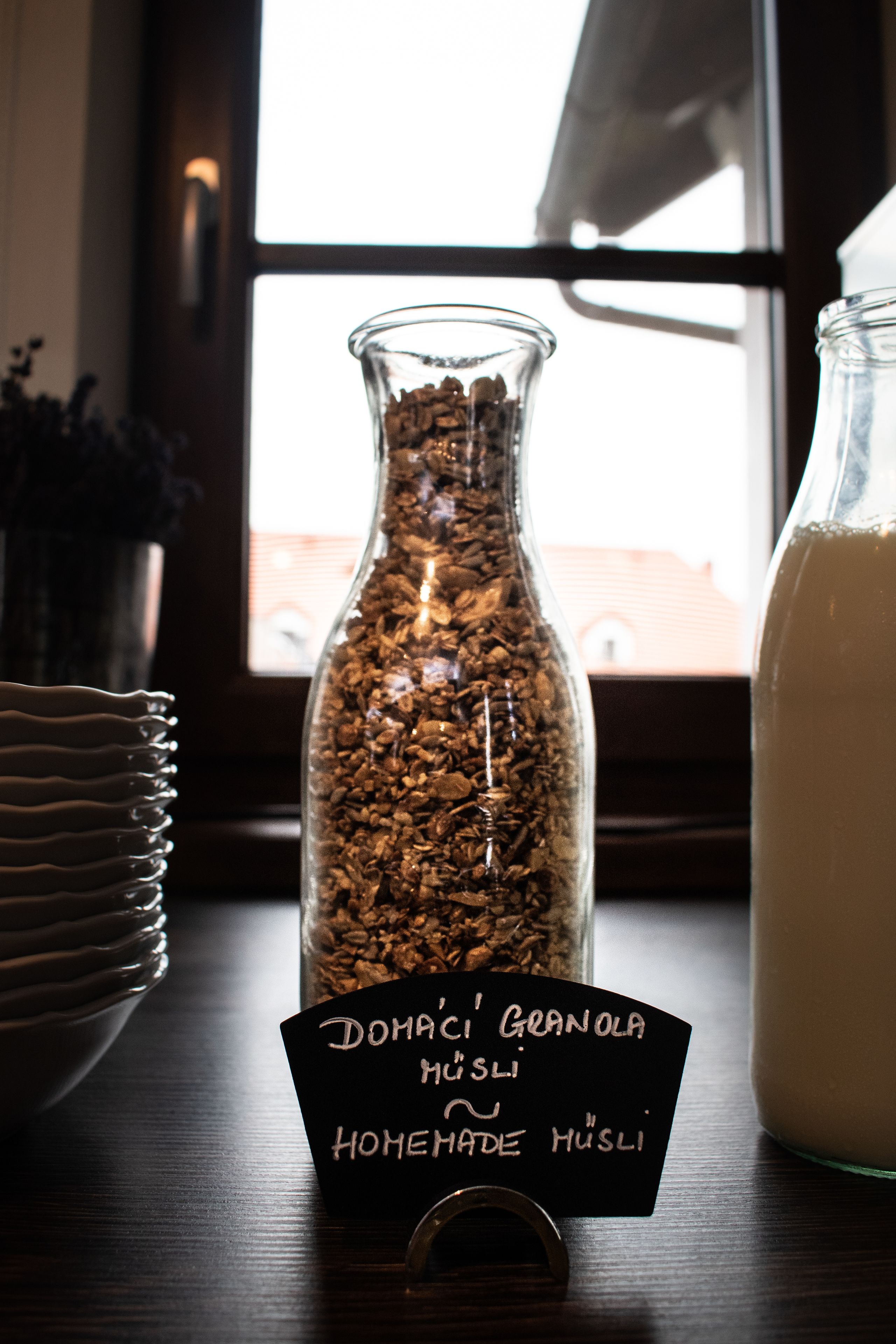 Ein Glasgefäß, gefüllt mit Granola, mit dem Aufdruck "Domaci' Granola Homemade Husli" auf einem Holztisch neben einem Glas Milch.