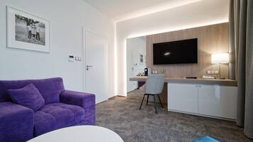 A modern hotel room with a purple sofa, a white round table, a kitchenette with a flat-screen TV, and a grey chair.