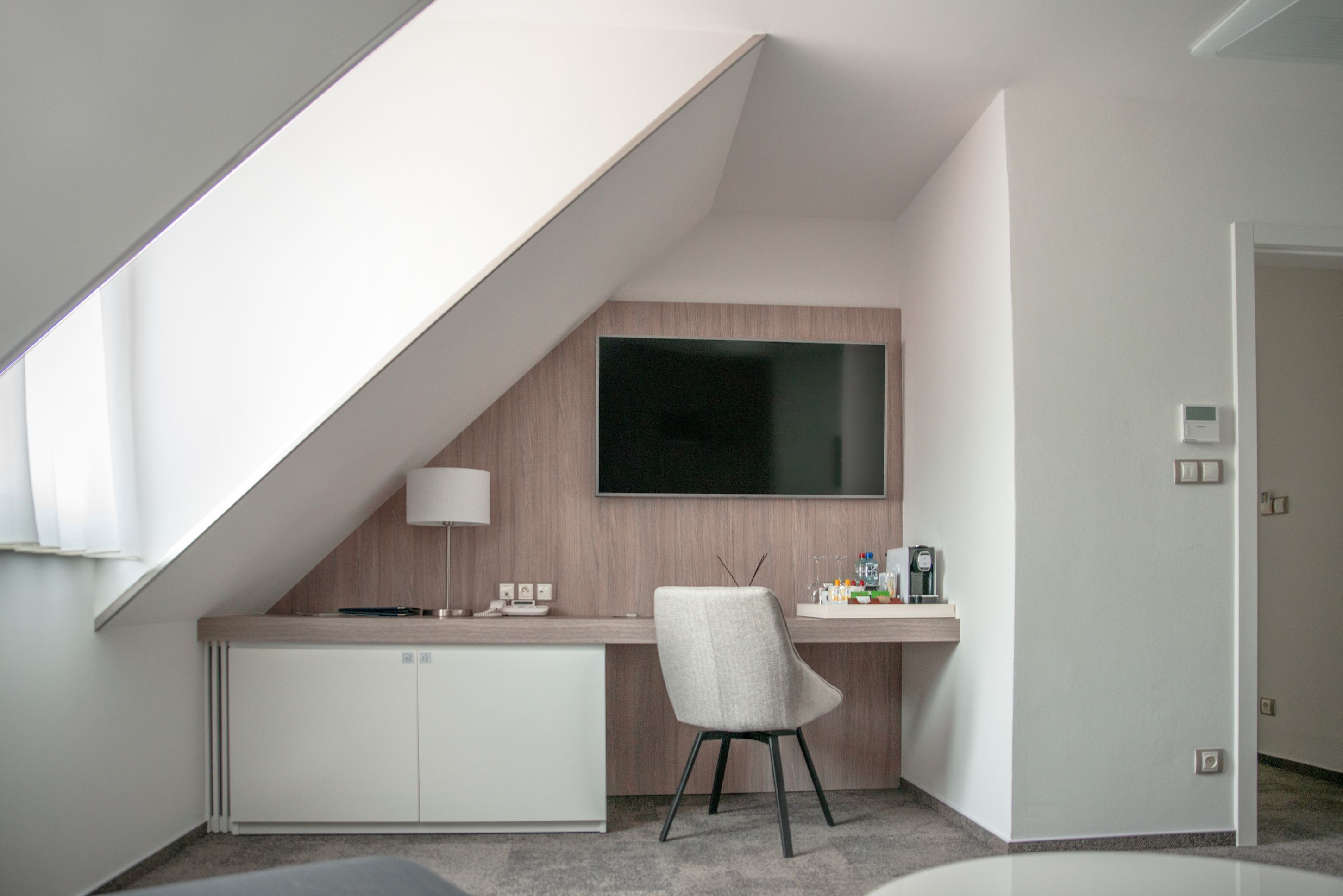 A modern, compact room with a sloped ceiling, a flat-screen TV mounted on the wall, a desk with a lamp, and a chair.