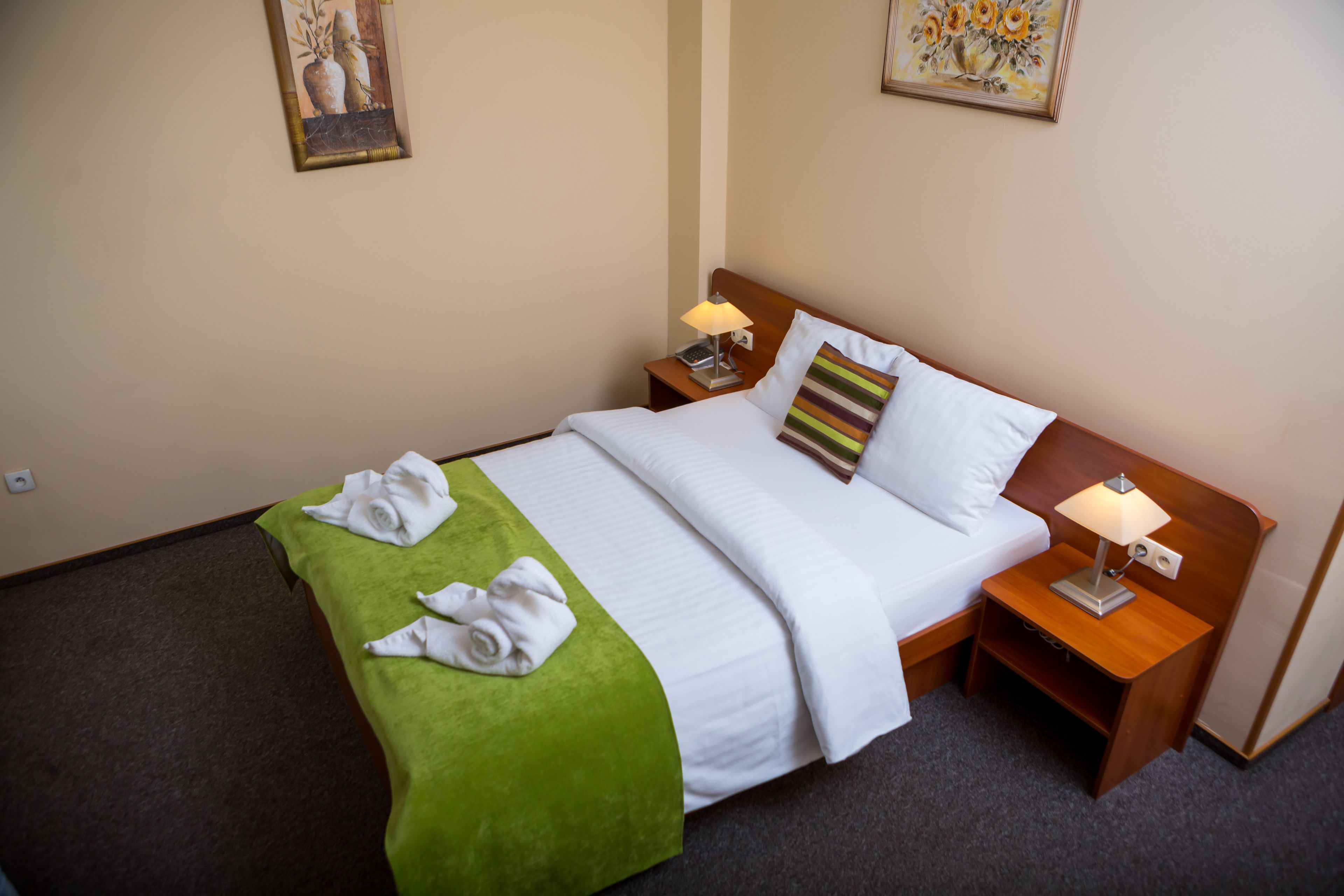 Cama bem arrumada com colcha verde, lençóis brancos e duas almofadas, incluindo uma listrada. Há duas mesas de cabeceira com abajures e uma pequena prateleira.