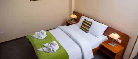 Cama bem arrumada com colcha verde, lençóis brancos e duas almofadas, incluindo uma listrada. Há duas mesas de cabeceira com abajures e uma pequena prateleira.