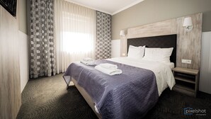 A hotel room with a large bed, a headboard with a light wood finish, a bedside table with a lamp, and a window with patterned curtains. - Hotel Classic (Rzeszów)