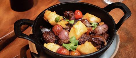 Plato en una sartén de hierro fundido sobre una mesa de madera, acompañado de una copa de vino y un vaso de agua.