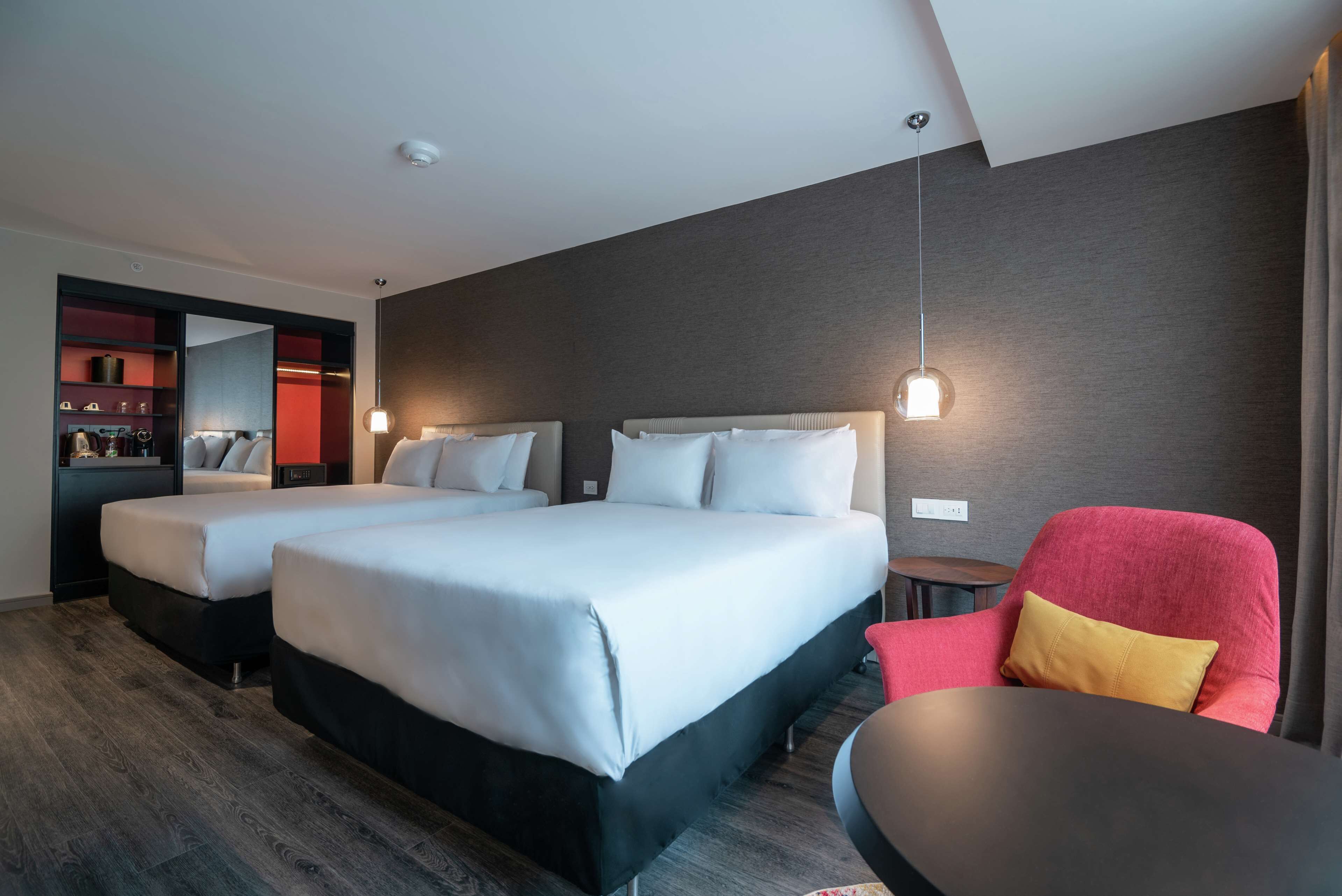 A hotel room with two beds, a red armchair, a small table, and a mirror.