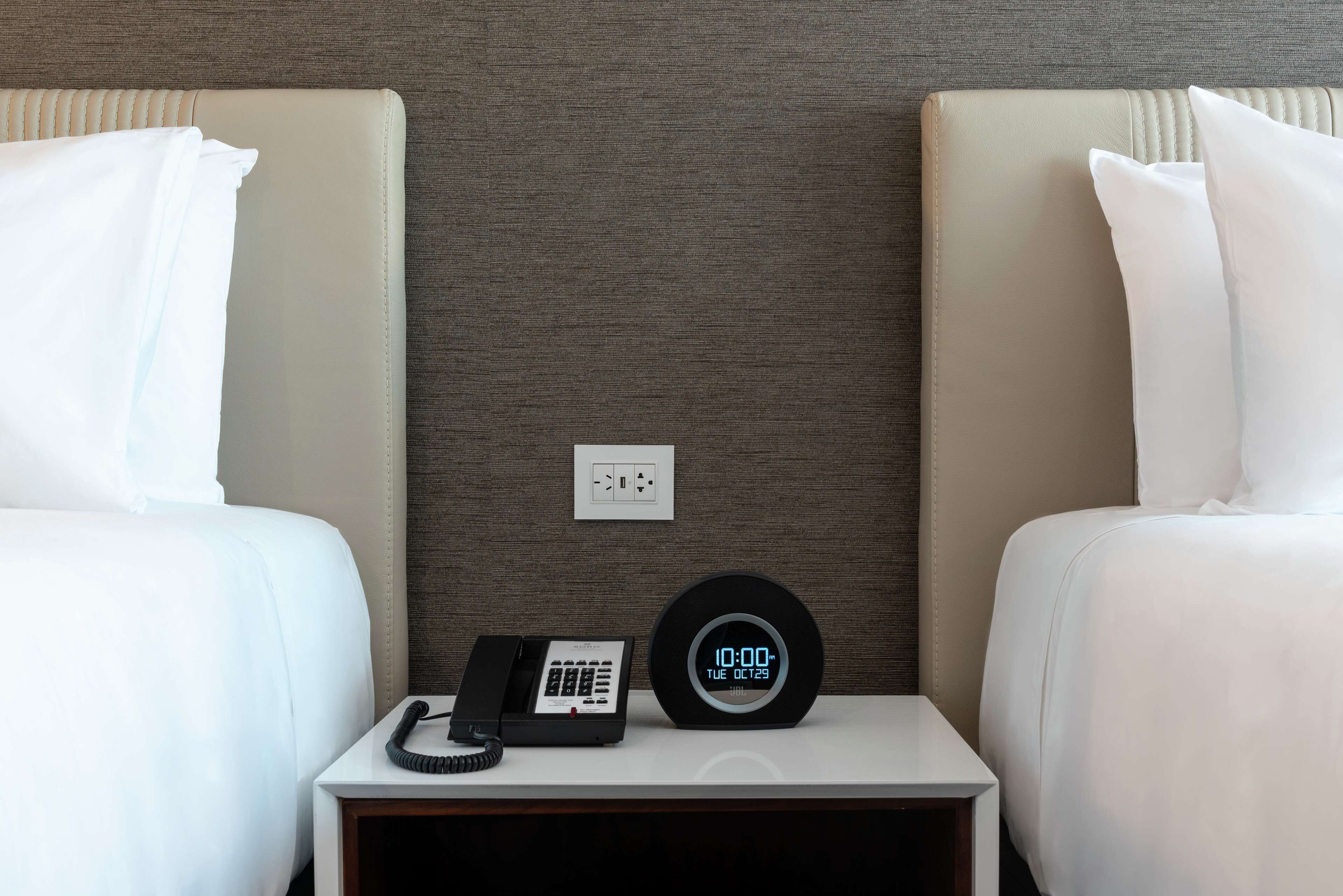 Habitación de hotel con dos camas, una mesita de noche con teléfono y reloj digital, y un enchufe de pared.