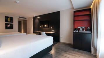 A modern hotel room with a large bed, a desk, a built-in kitchen area, and a red accent wall.