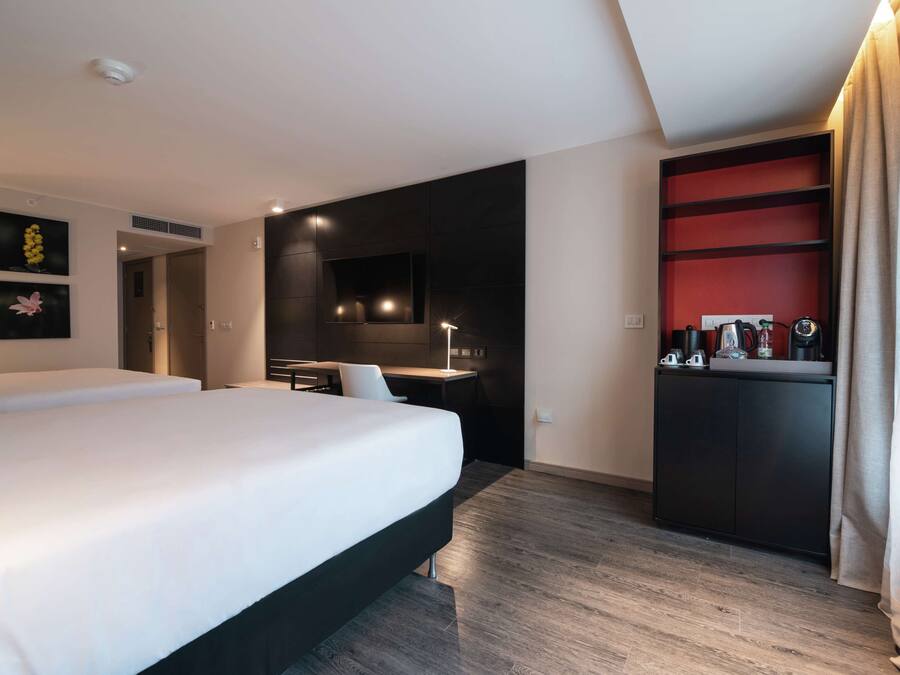A modern hotel room with a large bed, a desk, a built-in kitchen area, and a red accent wall.