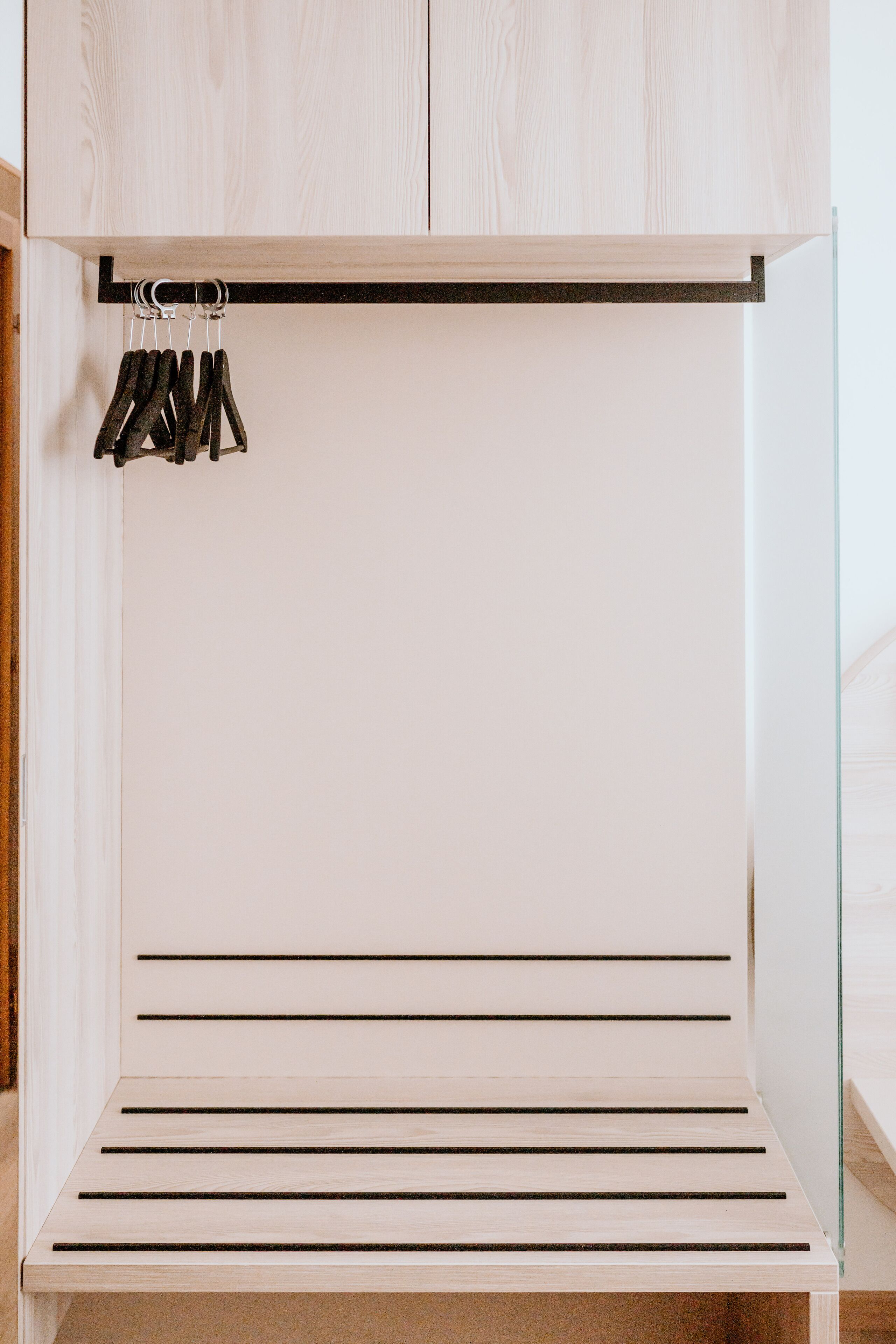A minimalist wooden wardrobe with a glass door, featuring multiple shelves and a hanging rod.