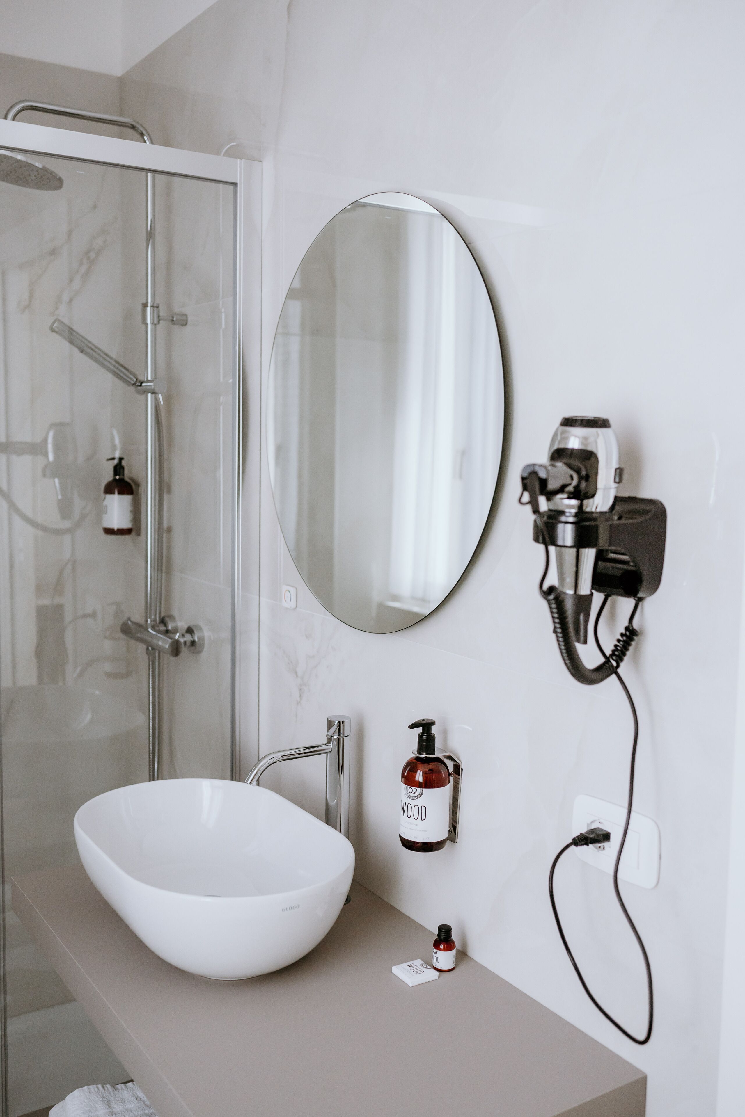 A bathroom with a white sink, a glass shower enclosure, a round mirror, and a hairdryer mounted on the wall.