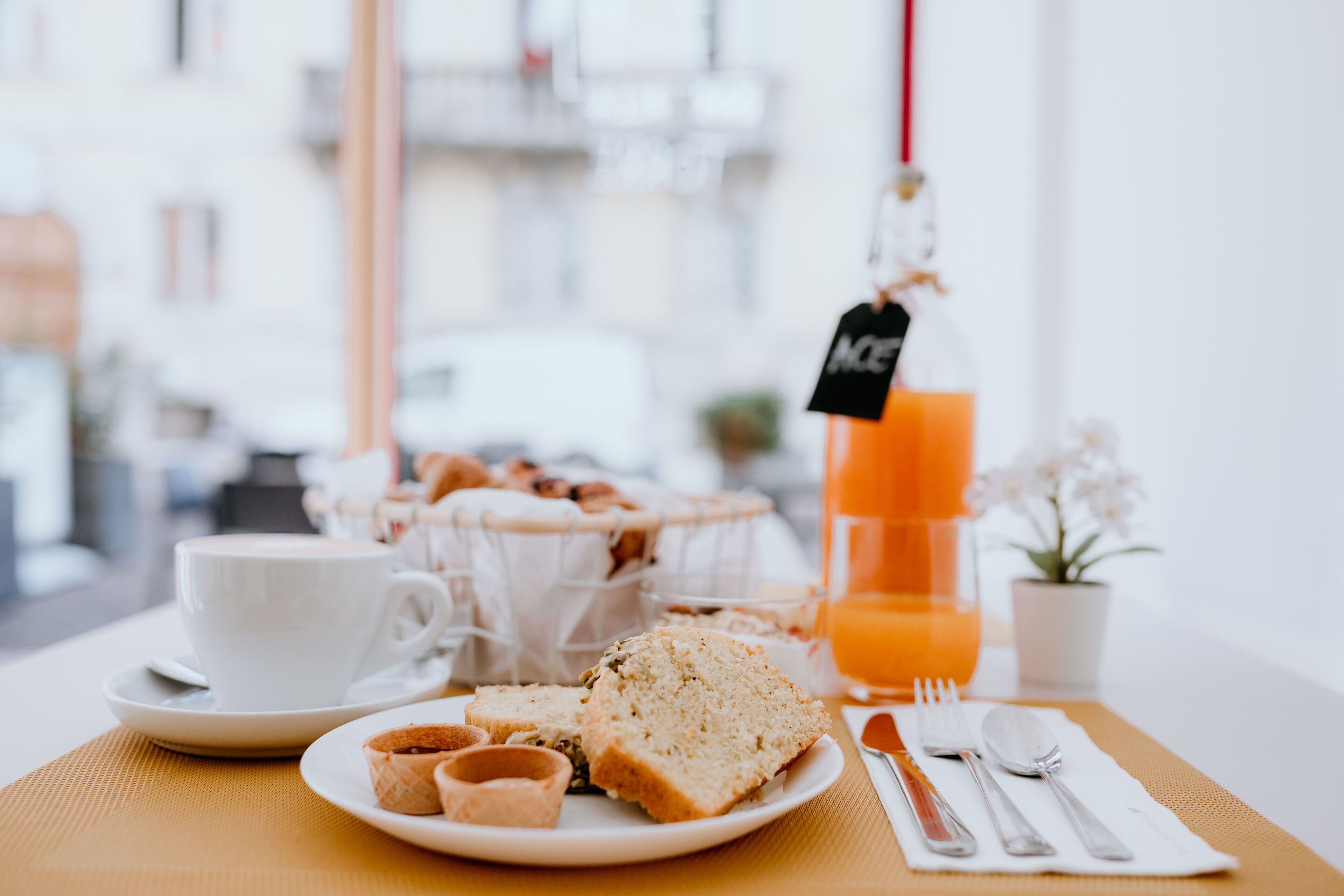 A breakfast setting with a white cup, a plate with bread and pastries, a glass of orange juice, and cutlery on a table.
