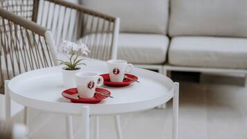 A white round table with two white cups and saucers, a small potted plant, and a wicker chair in a modern, minimalistic room with a white sofa in the background.