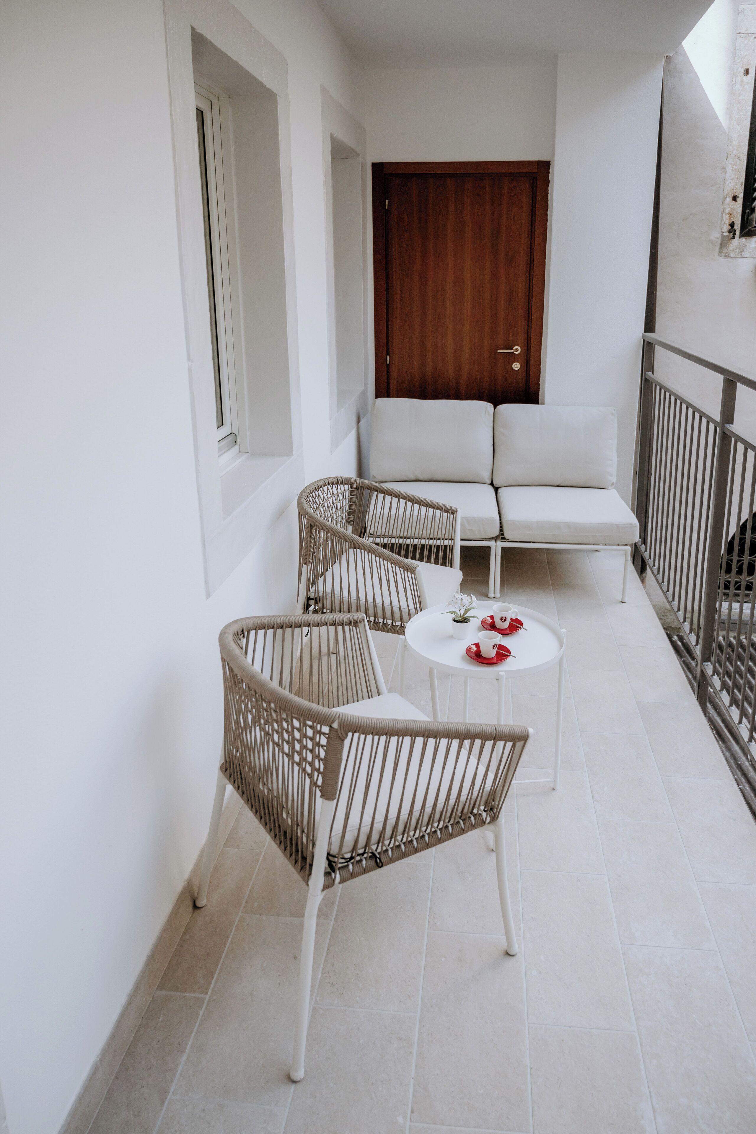 Un balcon avec un canapé blanc, des fauteuils en osier et une petite table sur laquelle sont posées des tasses rouges et blanches.
