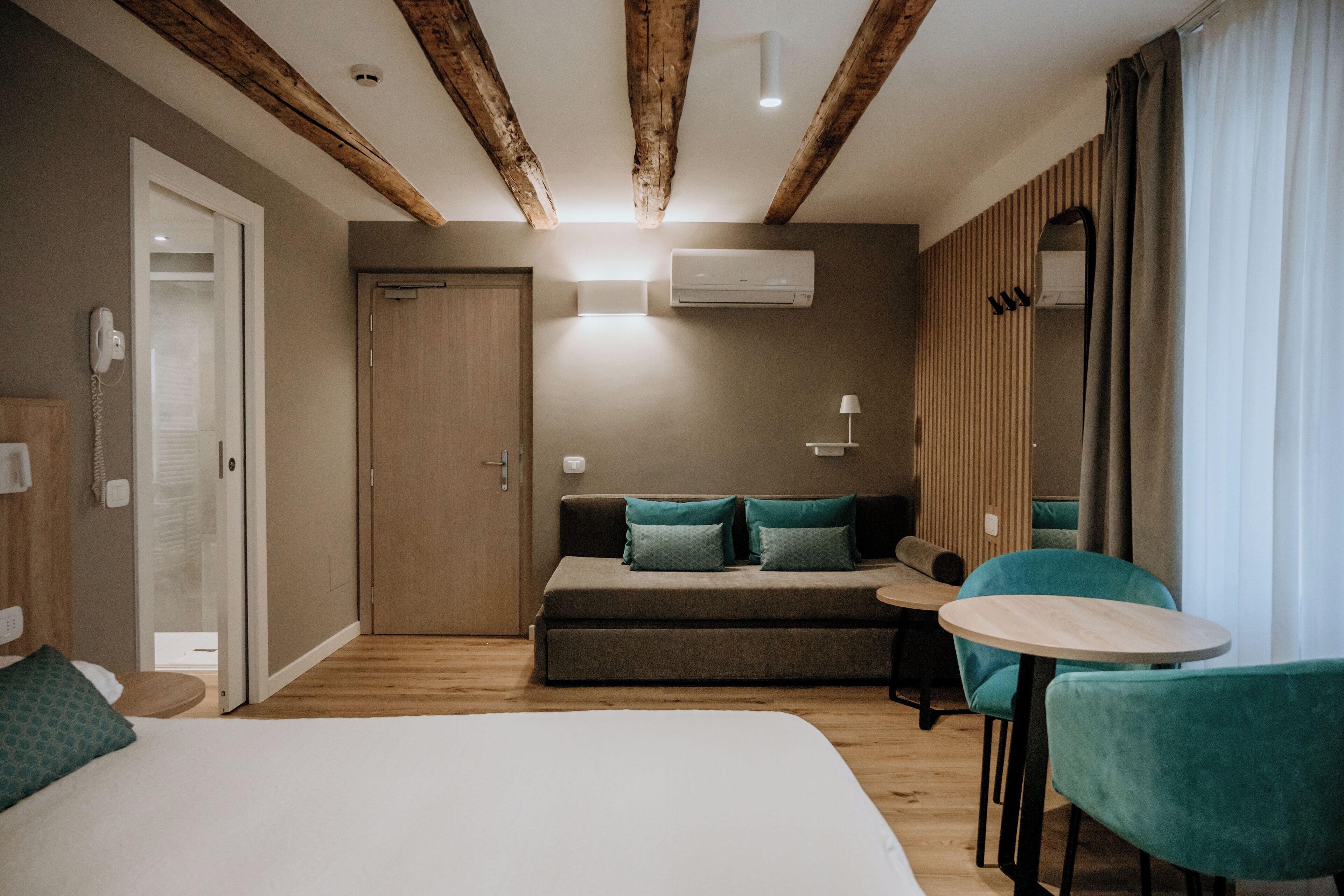 A modern hotel room with a large bed, a sofa, two chairs, a small table, wooden beams on the ceiling, and a door.