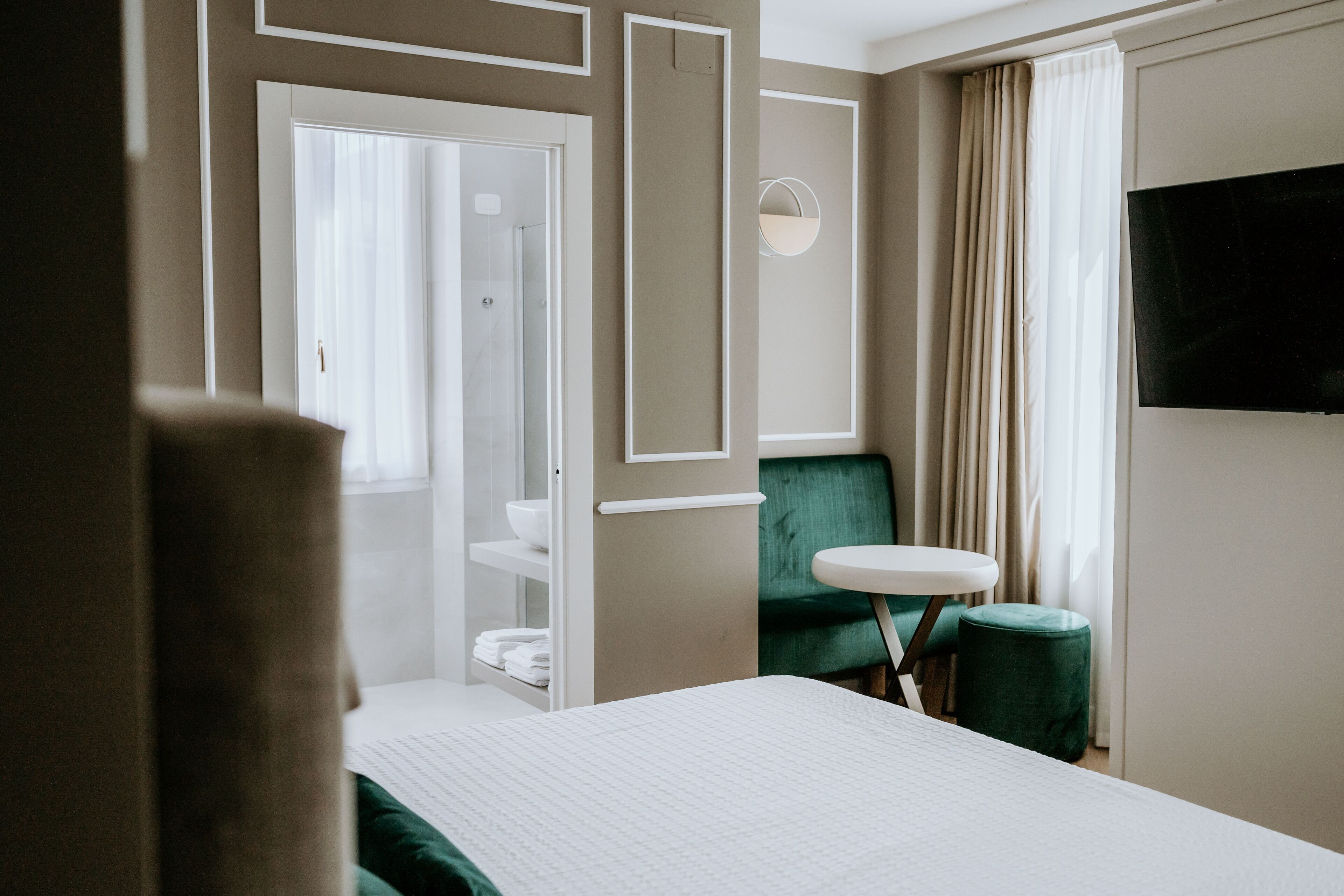 Habitación de hotel con cama, una mesita redonda pequeña, una silla verde, televisor montado en la pared y un baño visible a través de una puerta abierta.