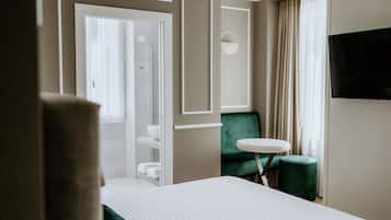 Habitación de hotel con cama, una mesita redonda pequeña, una silla verde, televisor montado en la pared y un baño visible a través de una puerta abierta.
