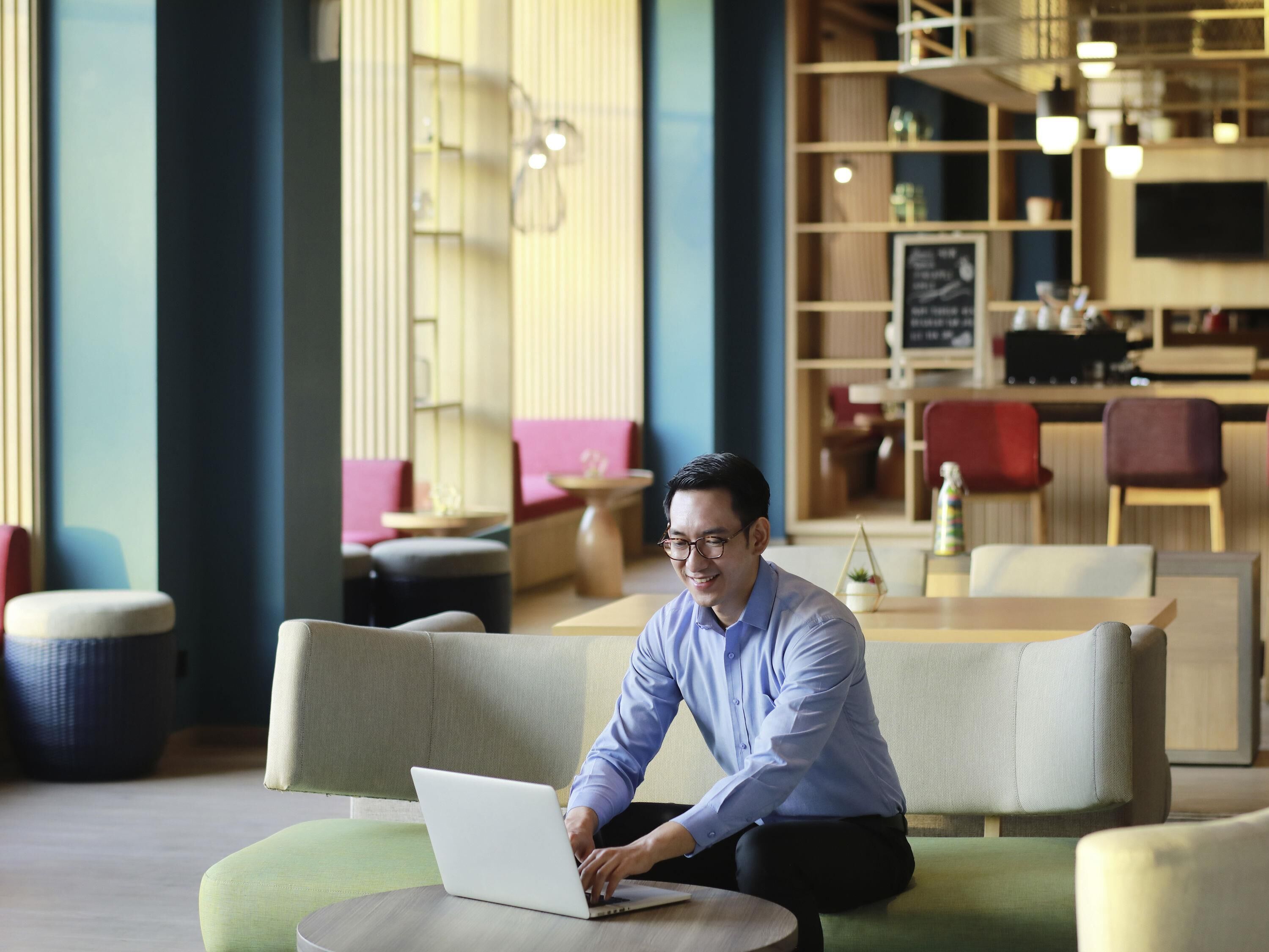A person sitting on a green sofa with a laptop, in a modern indoor setting with wooden shelves and a bar area in the background.