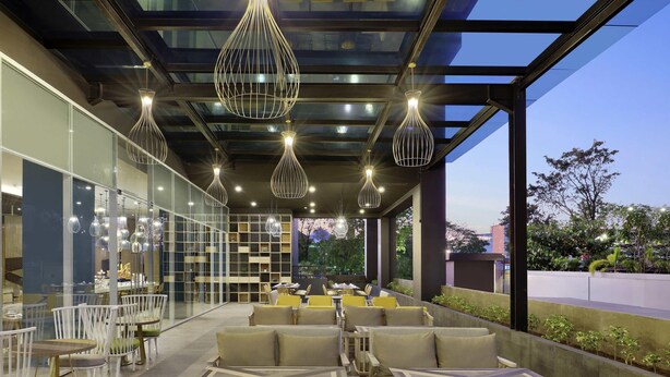 A modern outdoor seating area with multiple tables and chairs, under a glass ceiling with hanging lights.