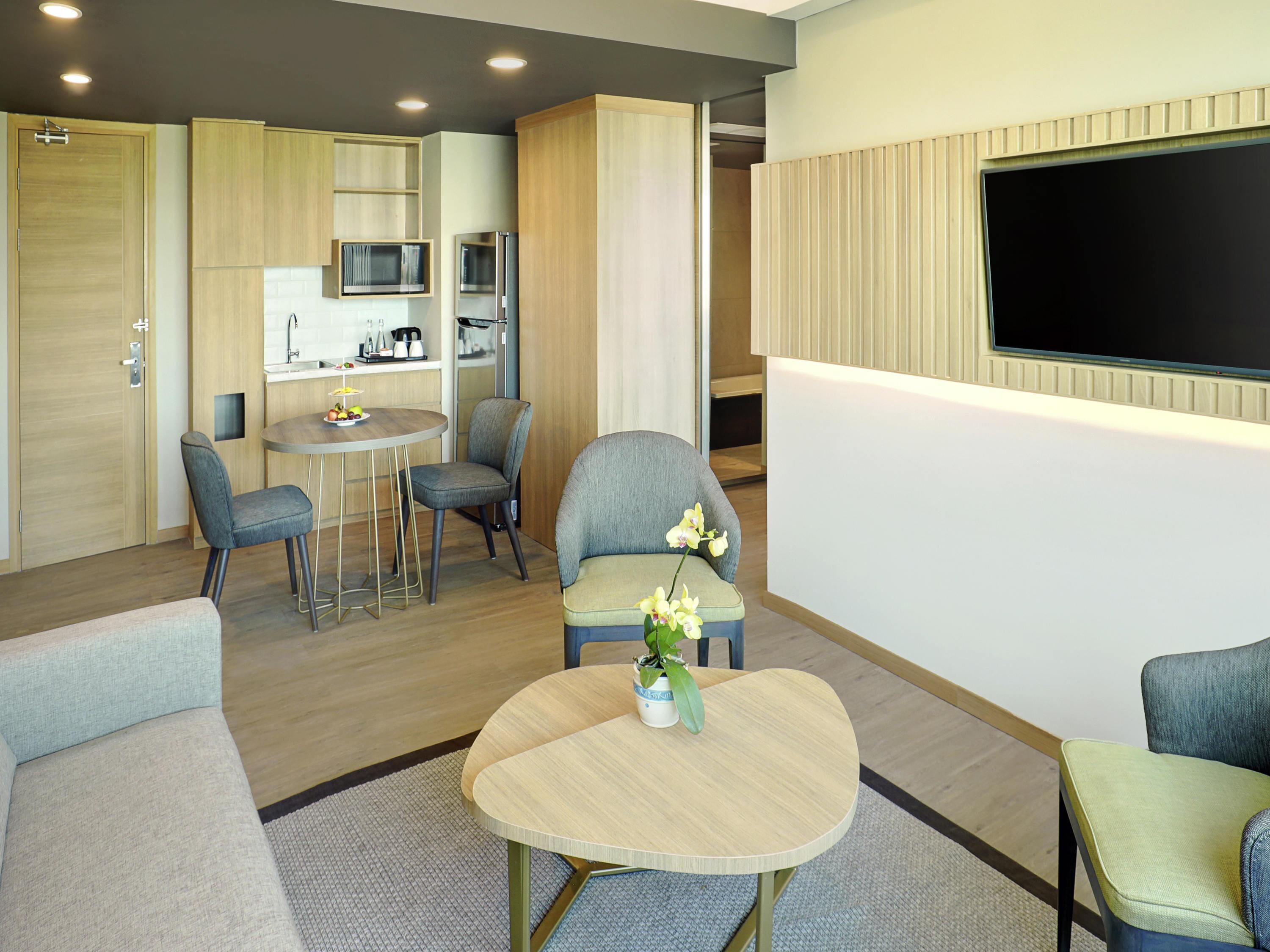 A modern hotel room with a dining area, kitchenette, and a flat-screen TV mounted on the wall.