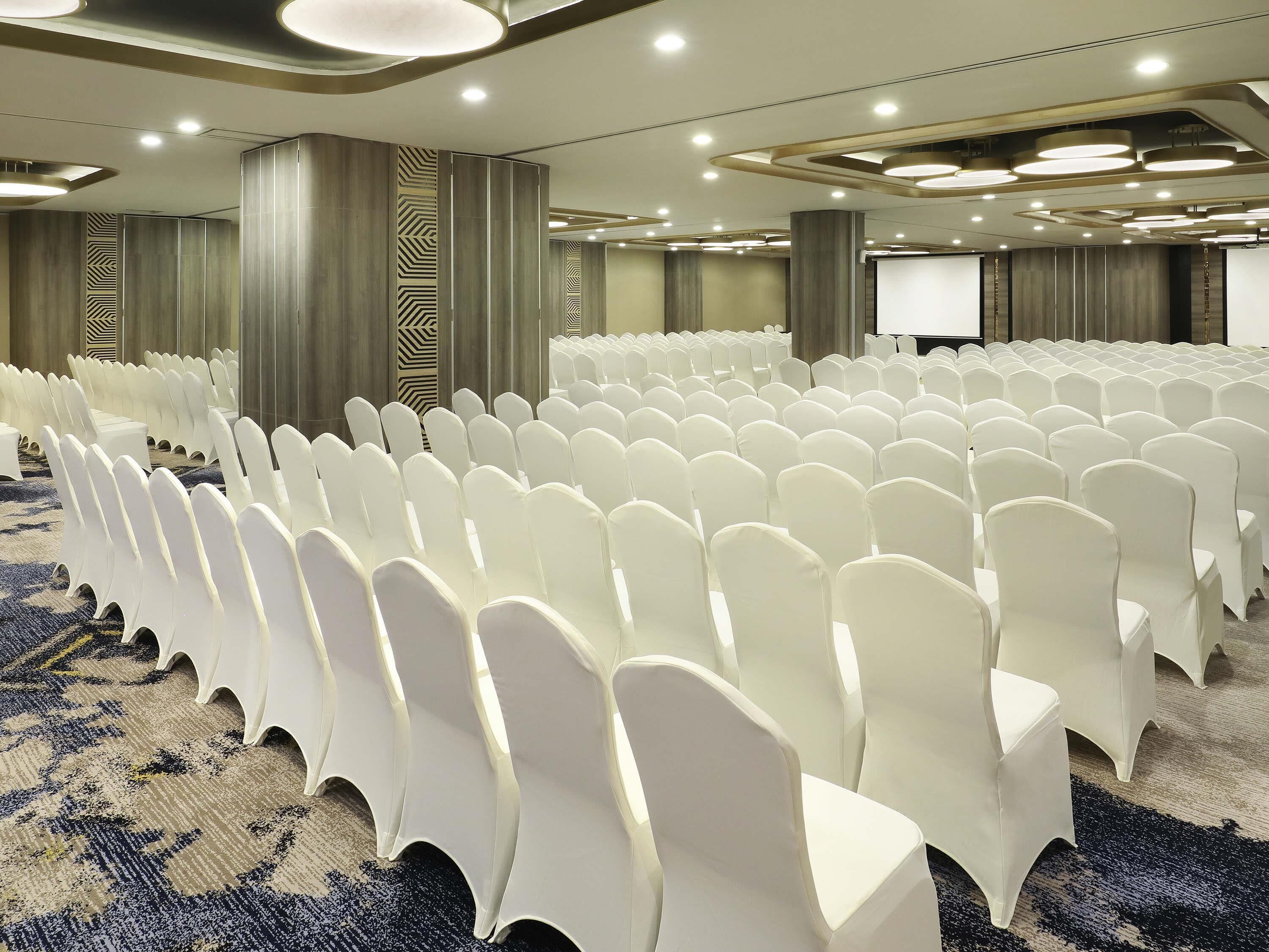 A conference room with rows of white chairs, a projector screen, and a carpeted floor.