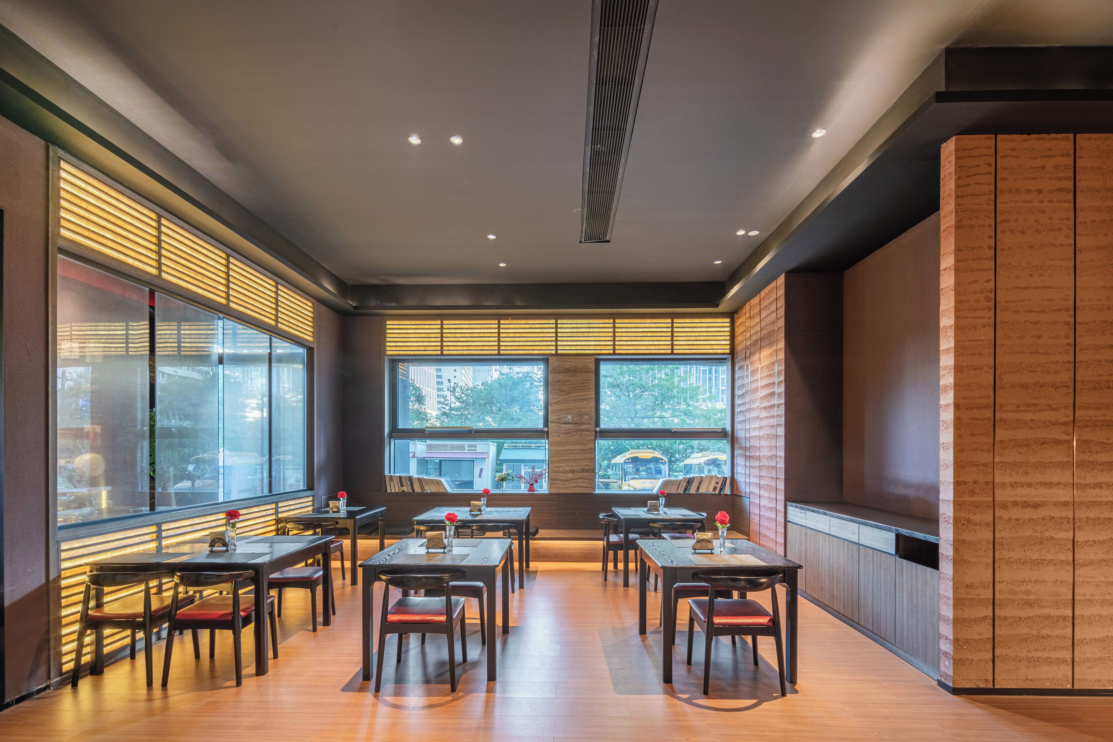 A modern restaurant with wooden tables and chairs, large windows with blinds, and a view of the city.