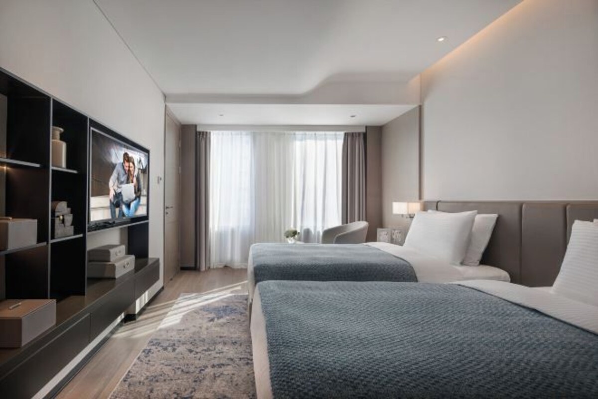 Una habitación de hotel moderna con dos camas, un armario amplio, un televisor de pantalla plana y un ventanal con cortinas.