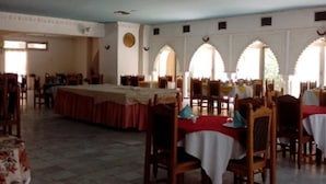 Une salle à manger spacieuse, dotée de hauts plafonds et de fenêtres en arc de cercle. Les tables sont dressées avec des nappes blanches et des chaises.