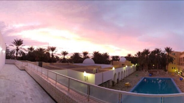 Une piscine d’hôtel avec vue sur le coucher de soleil, des palmiers et un ciel dégagé.