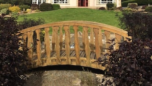 Een groot, twee verdiepingen tellend huis met een rode deur en meerdere ramen, omgeven door een verzorgde tuin en een houten brug.