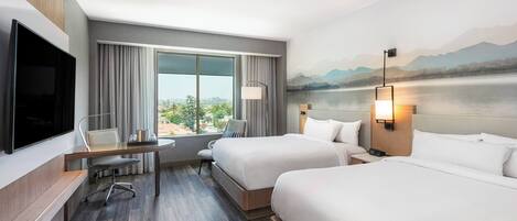 A hotel room with two beds, a desk with a chair, a TV, and a large window with curtains.