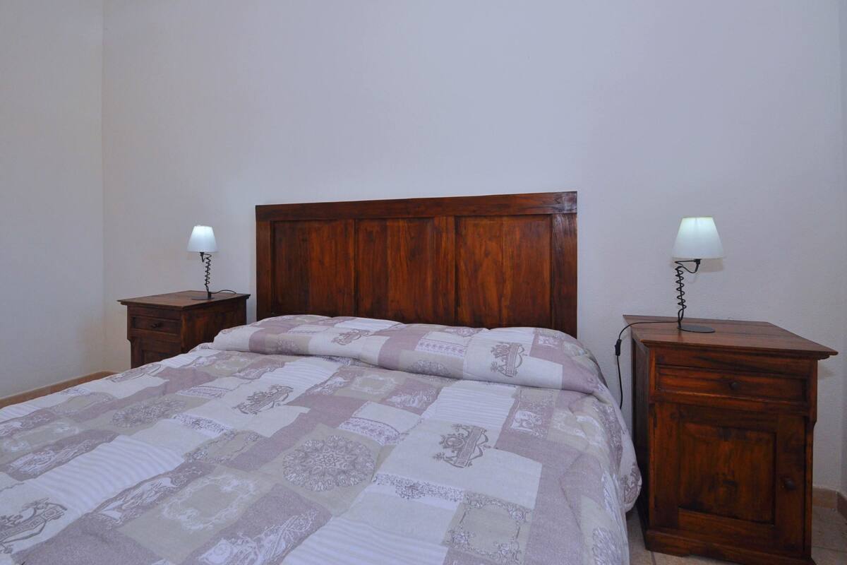 Un dormitorio con cabecera de madera, colcha acolchada, dos mesitas de noche con lámparas y una mesita auxiliar.