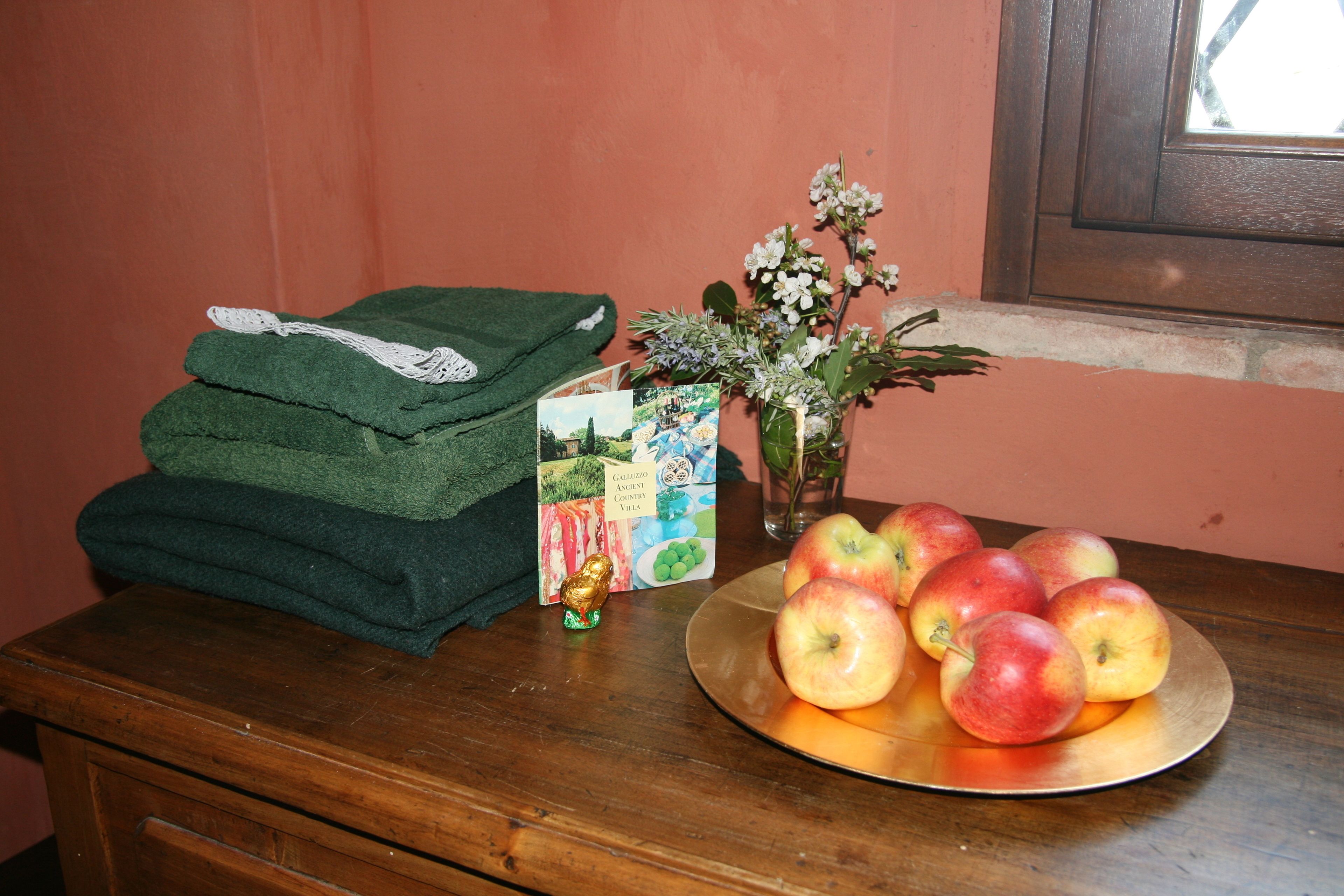 A wooden table with a stack of folded green towels, a small decorative item, a card, a vase with flowers, and a plate with apples.