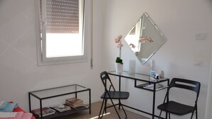 Une petite chambre moderne, équipée d’un lit, d’un bureau, d’une chaise, d’un miroir et d’une fenêtre avec des stores.