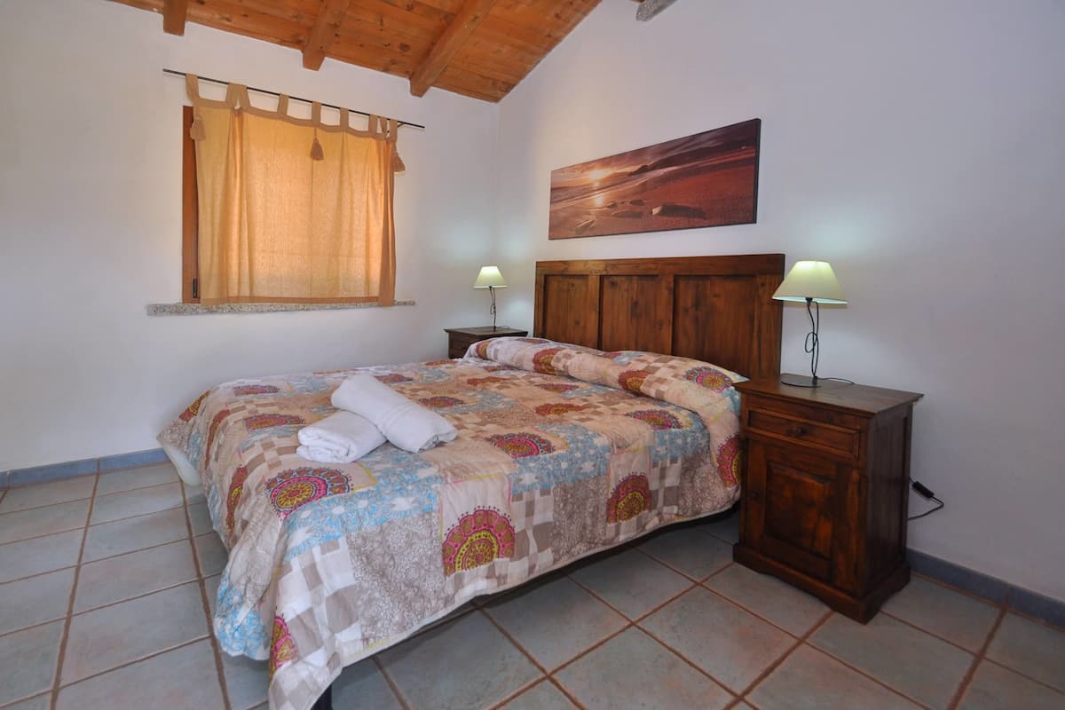 A bedroom with a wooden bed, two bedside tables with lamps, a window with curtains, and a painting on the wall.