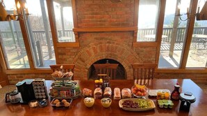A dining table with various food items and a coffee maker in a room with a brick fireplace and large windows. - Bent Mountain Lodge (Copper Hill)