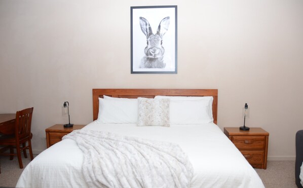Una cama bien hecha con edredón blanco y manta, flanqueada por dos mesitas de noche de madera con lámparas, una silla de madera y un cuadro enmarcado de un conejo en la pared.