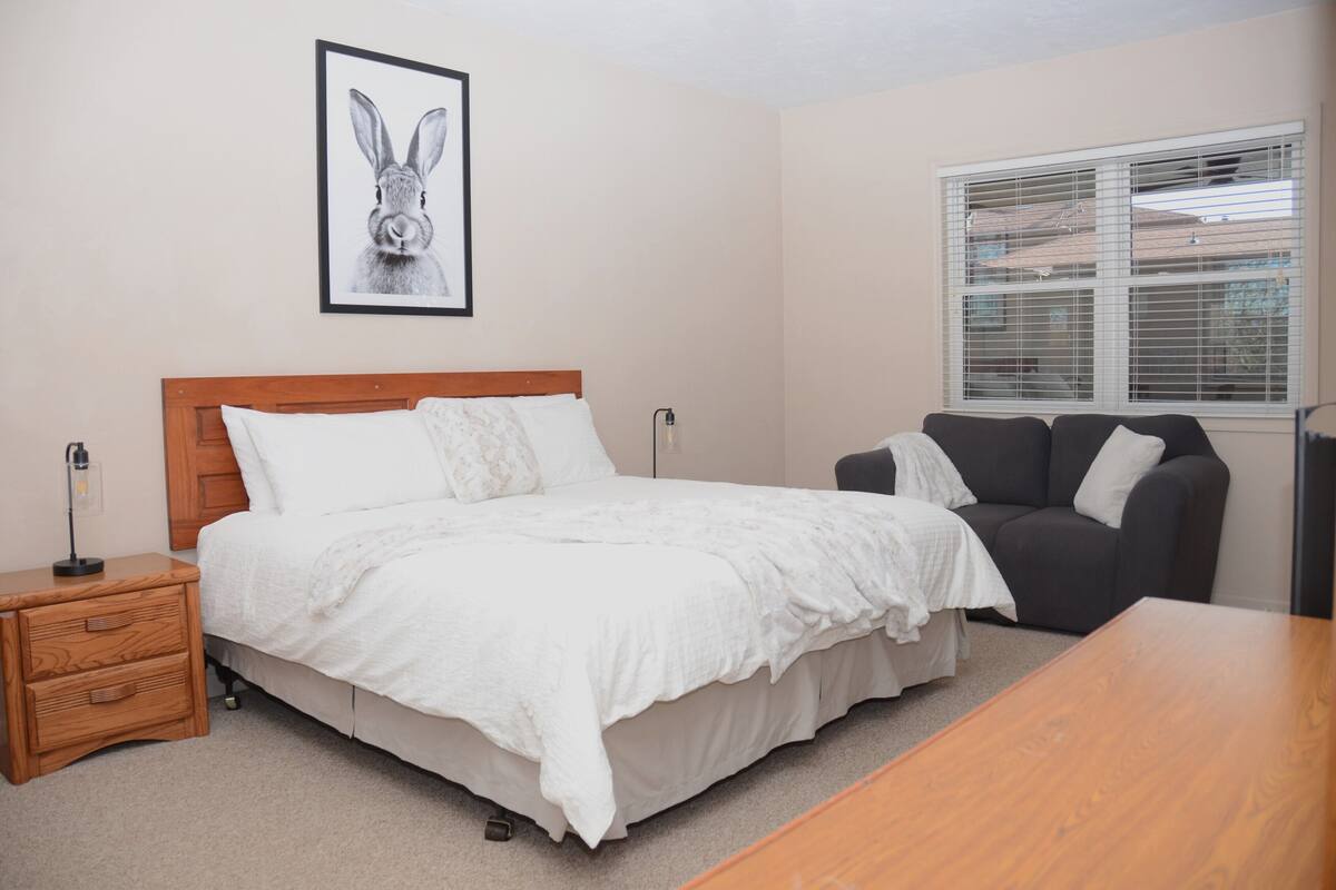 Een slaapkamer met een houten bed, een nachtkastje, een grijze bank en een raam met jaloezieën.