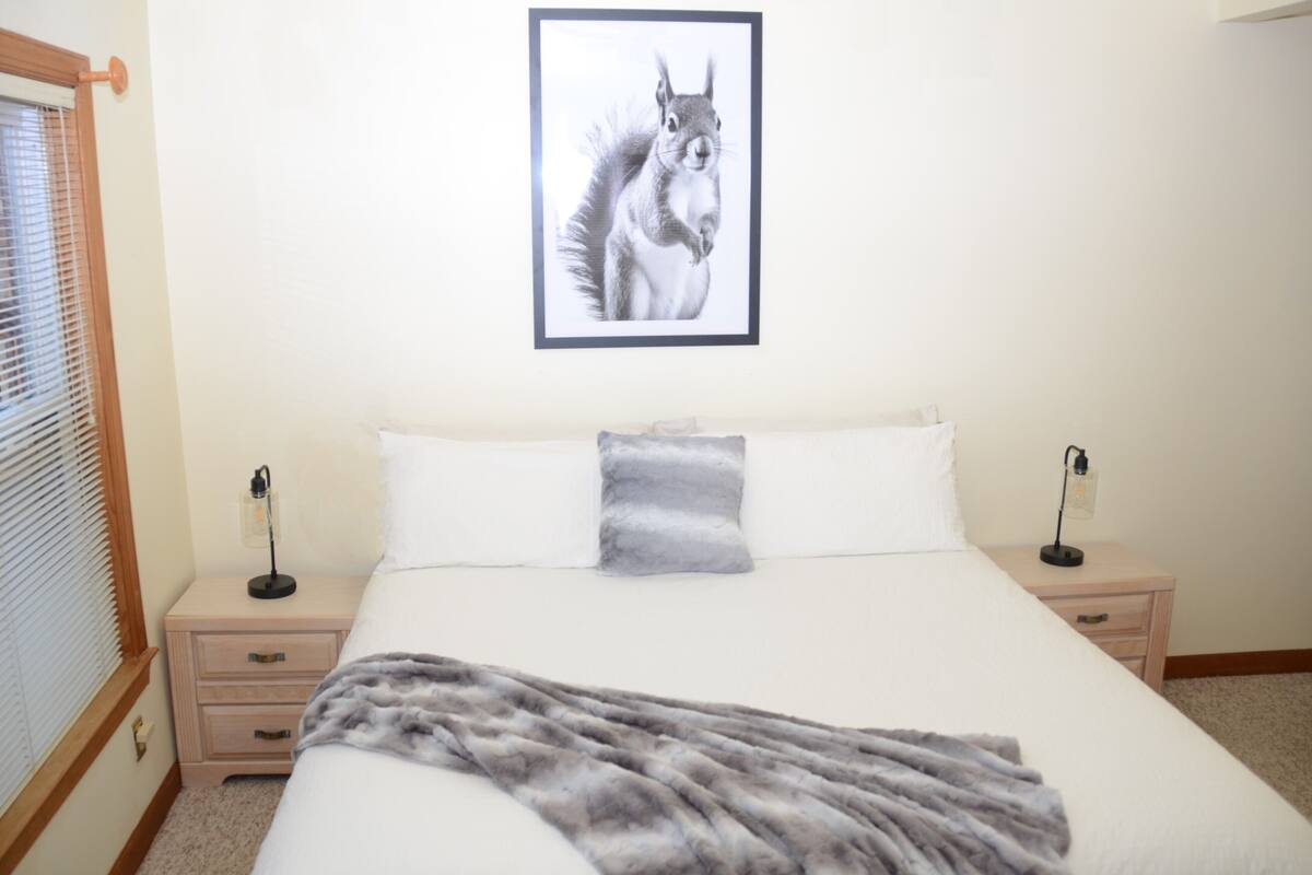 Een slaapkamer met een bed, twee nachtkastjes, een lijst met een foto aan de muur, en een raam met zonwering.