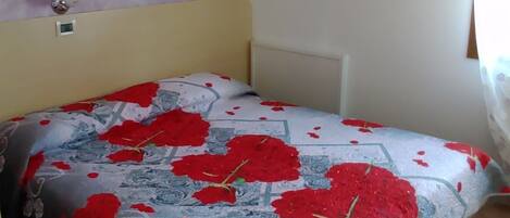 Un dormitorio con cama, una silla amarilla pequeña, un crucifijo en la pared y una ventana con cortinas.