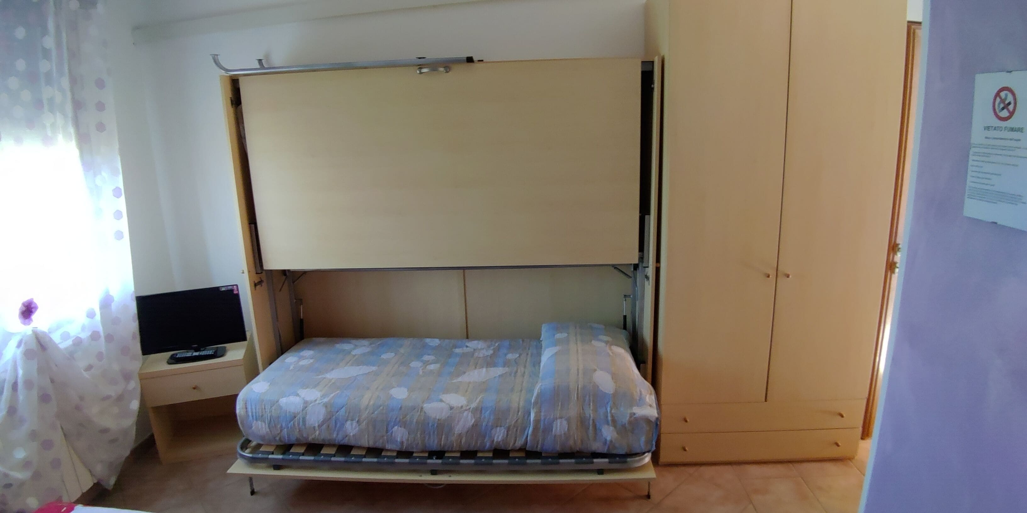Quarto com uma cama de solteiro, uma escrivaninha com computador e uma janela com cortinas.