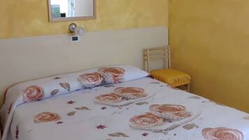 Un dormitorio con colcha estampada, mesita de noche, una silla, un espejo montado en la pared y una cruz.