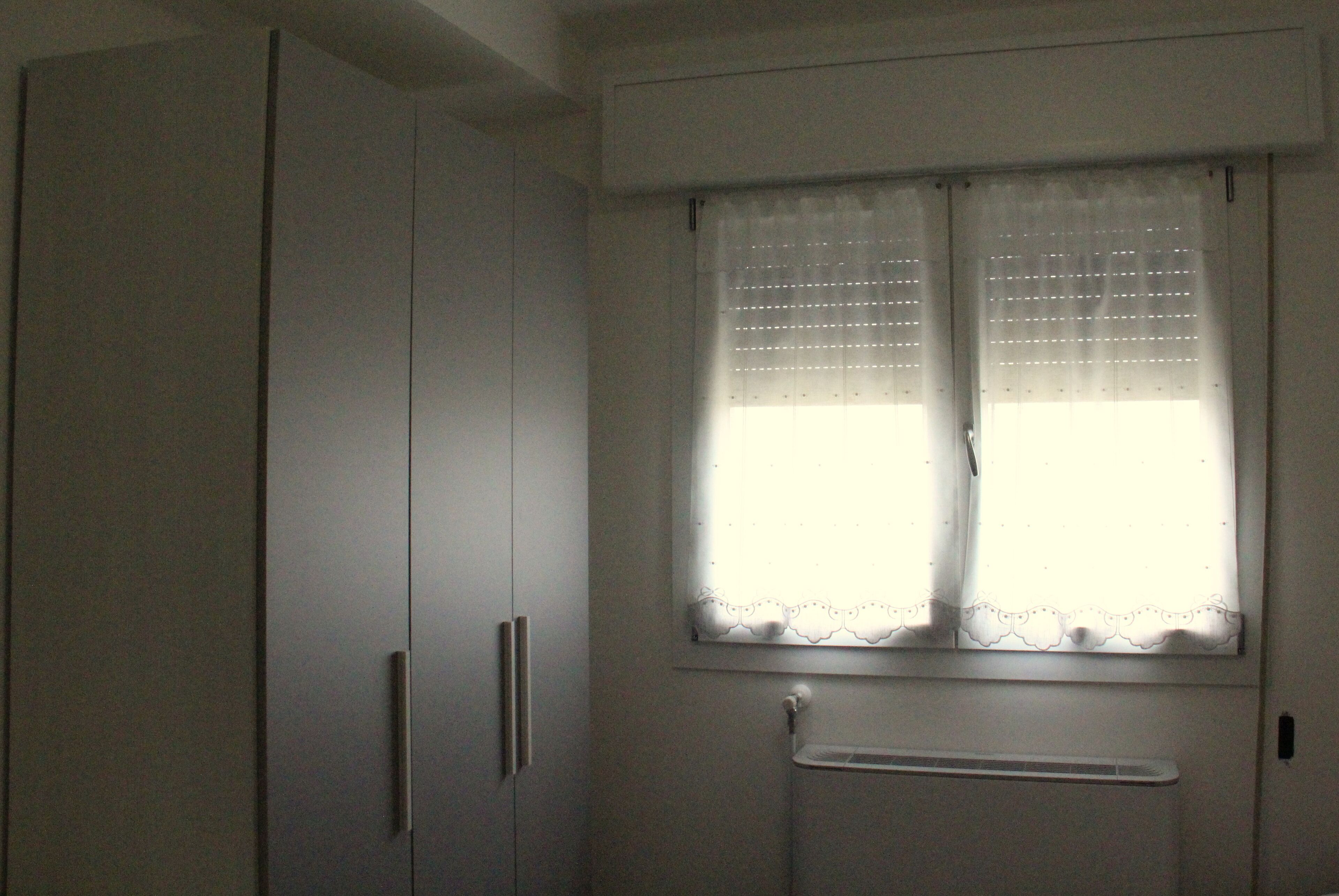 A room with a large wardrobe, a window with a lace curtain, and a radiator.